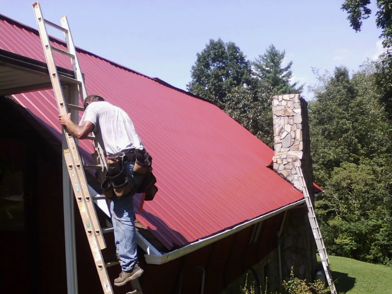 Roofer on a ladder beside a red metal roof and stone chimney — Soffit & Fascia Repair by Roofing Brothers in Loomis